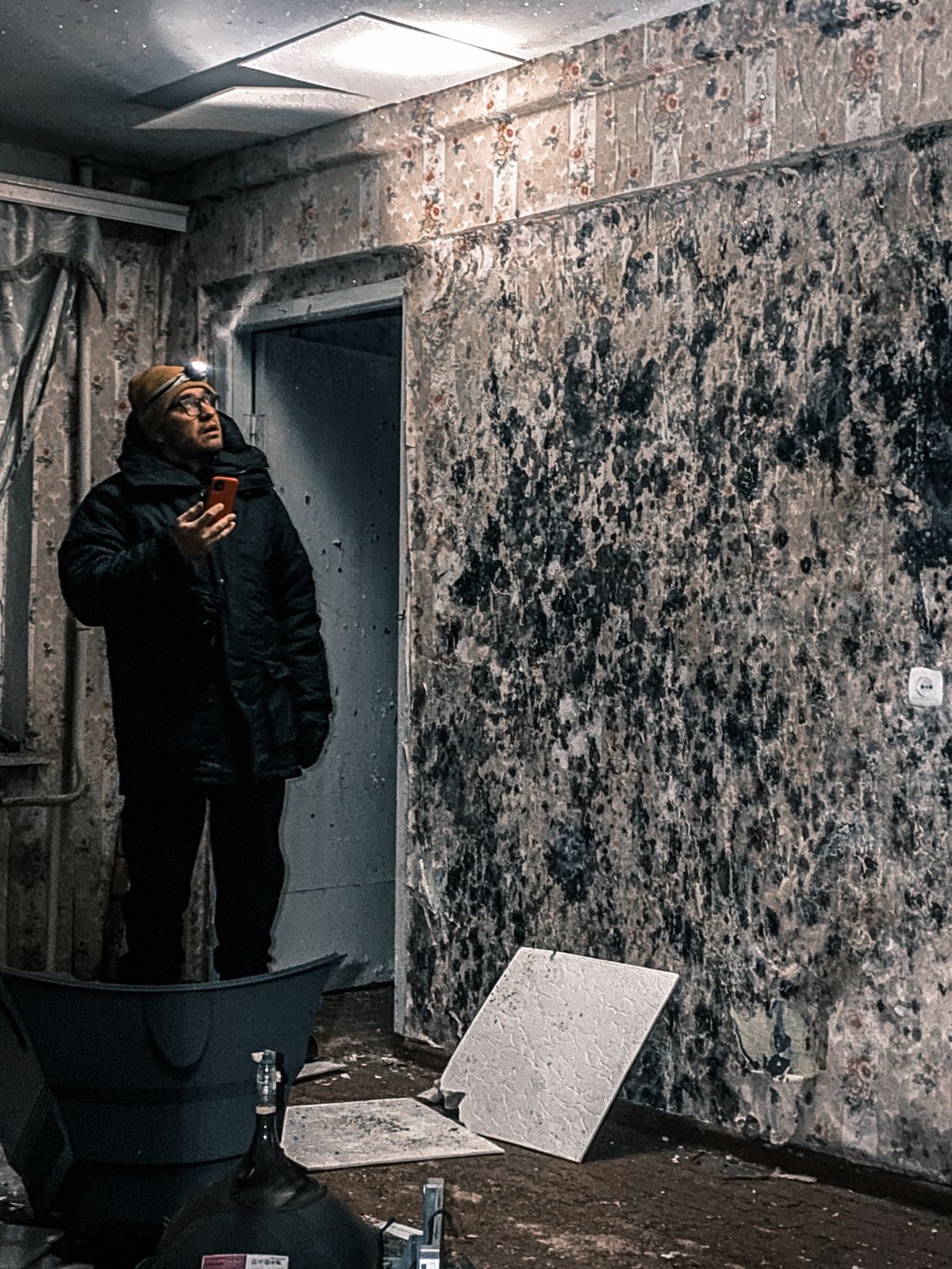 Inspector with a headlamp examining the deteriorated walls and ceiling tiles of an older building